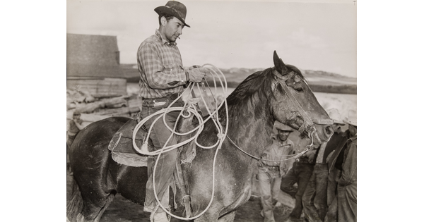 A black-and-white photograph of a light-skinned man in a brimmed hat holding a lasso on horseback.