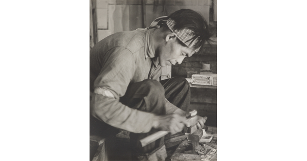 A black-and-white photograph of a seated Native American man bent over an anvil in a workshop.
