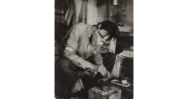 A black-and-white photograph of a seated Native American man bent over an anvil in a workshop.