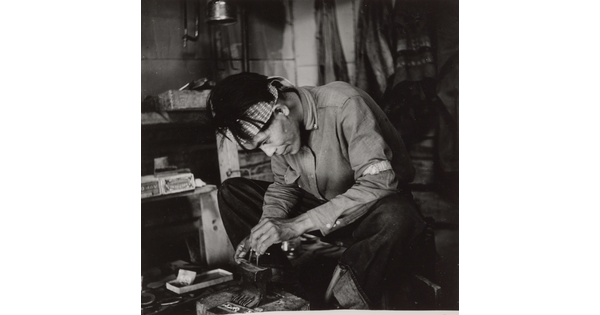 A black-and-white photograph of a seated Native American man bent over an anvil in a workshop.