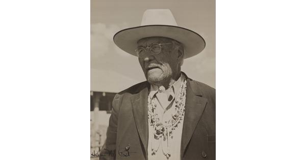 A black-and-white photograph of a Native American man wearing a wide-brimmed hat, glasses, a suit jacket and several necklaces.