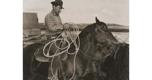 A black-and-white photograph of a light-skinned man in a brimmed hat holding a lasso on horseback.