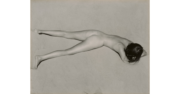 A black-and-white photograph of a nude, fair-skinned woman lying facedown in the sand with her head resting on her crossed arms.