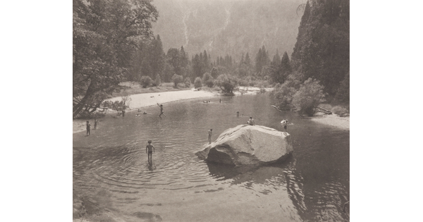 A black-and-white photograph of people swimming in a pond, a large stone in the center, surrounded by trees and mountains.