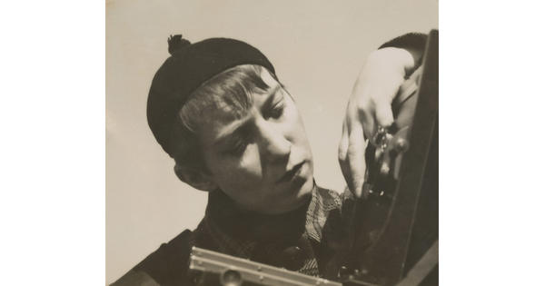 A black-and-white photograph of a White woman with short hair, wearing a beanie hat, manipulating a large format camera.