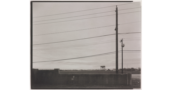 A black-and-white photograph of low brick wall beneath powerlines and utility poles and a water tower in the distance.