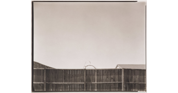 A black-and-white photograph of a wood privacy fence; the top of a pool slide and a roof are seen just above it.
