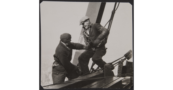 A black-and-white photograph of two men teasing one another as they balance on steel girders and cables high above the ground.