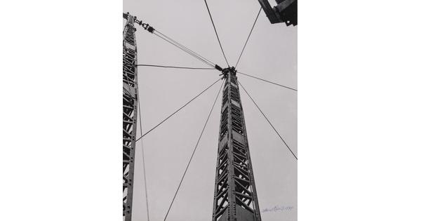 A black-and-white photograph of a steel derrick rising toward the sky with eight cables radiating from the top.