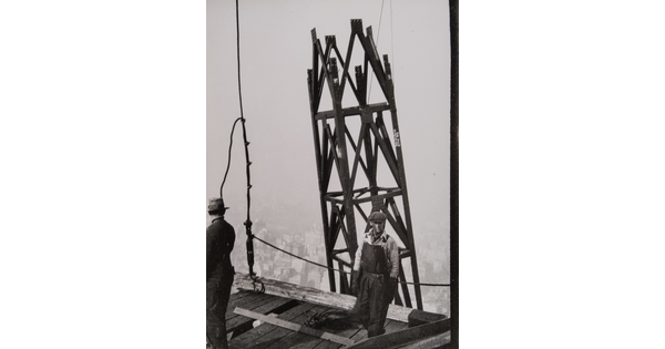 A black-and-white photograph of two men on a wooden platform of a construction site and a steel tower structure next to them.