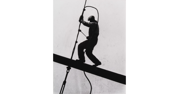 A black-and-white photograph of a man suspended precariously on an I-beam working with a tool on a cable, all in silhouette.