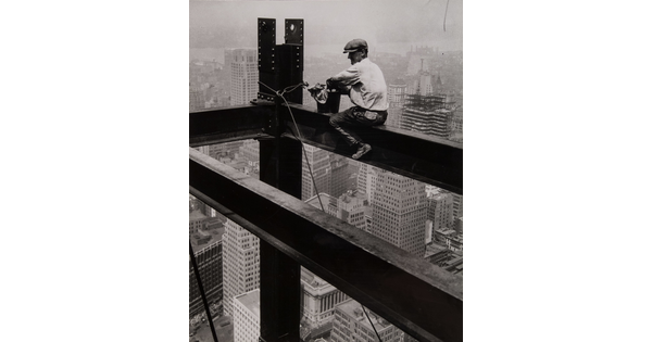A black-and-white photograph of a man wearing a driving cap sitting on a steel beam high above a large city.
