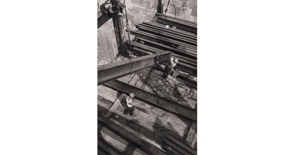 A black-and-white photograph from above of two men guiding a steel girder onto a platform high over a city.