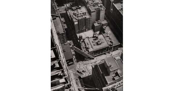 A black-and-white photograph looking down on the buildings of a city and several steel girders suspended by cables in mid-air.