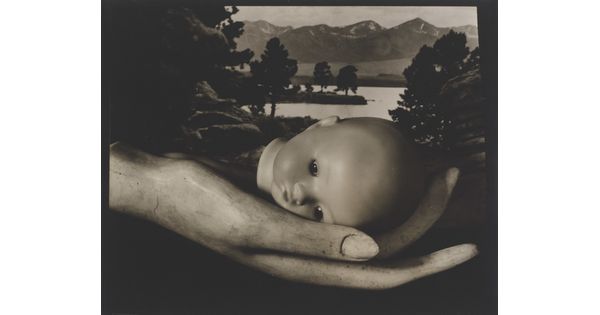 A black-and-white photograph of a mannequin hand holding a baby doll head in front of a landscape backdrop.