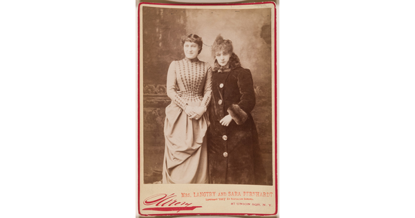 A sepia-toned studio portrait photograph of two elegantly dressed women in 1880s clothing above a bold red signature by the photographer.