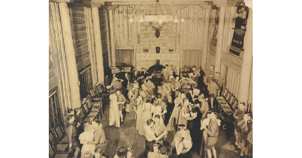 A sepia-toned photograph of a large group of people dancing in a large room, as viewed from above.