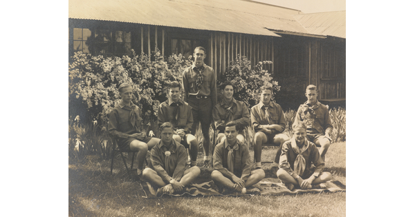 A sepia-toned photograph of a group of light-skinned young men in uniforms posed outside a building.