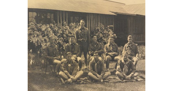 A sepia-toned photograph of a group of light-skinned young men in uniforms posed outside a building.