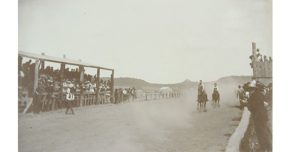 A black-and-white photograph of people on running horses coming around a bend in a dirt racetrack with spectators in stands on either side.