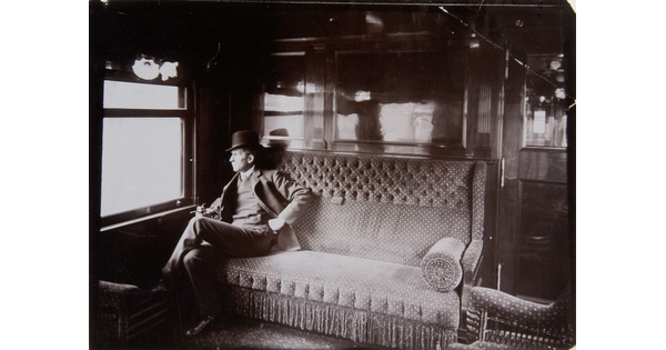 A black-and-white photograph of a White man wearing a top hat sitting on a couch in a railroad car, gazing out the window.