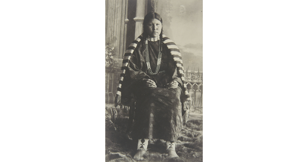 A black-and-white photograph of a seated Native American woman in traditional clothing with a striped blanket over her shoulders.