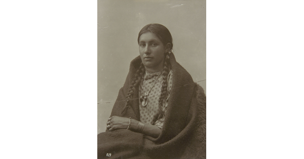 A black-and-white photograph of a seated Native American woman in traditional clothing with her hair in braids and a blanket around her shoulders.