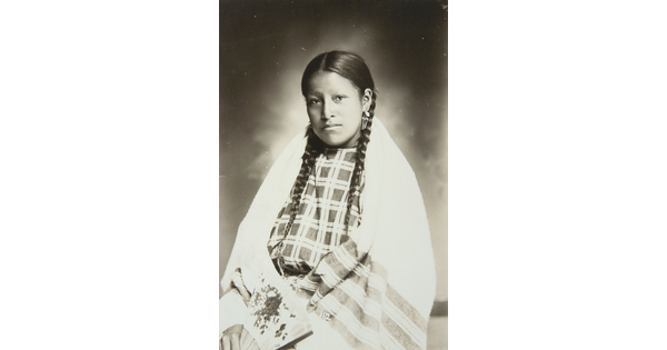A formal black-and-white portrait photograph of a seated Native American girl, hair in braids, wearing cultural dress, and holding a fan.