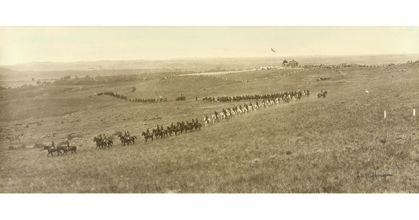 A black-and-white photograph of a grassy plain with a line of cavalry on horseback travelling through the landscape.