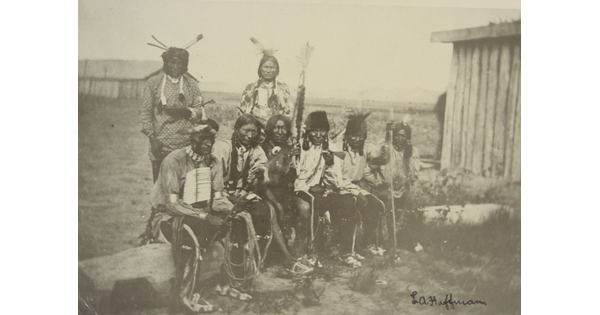 A black-and-white photograph of six Native Americans sitting on a log and two standing behind them, all wearing traditional clothing.