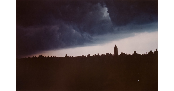 A color photograph of dark blue-gray storm clouds over the silhouette of a city skyline.