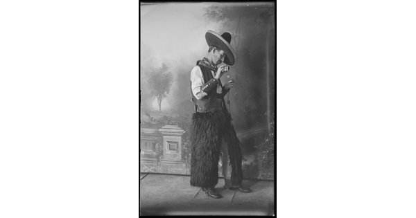A black-and-white, studio portrait image of a man standing in front of a backdrop wearing a sombrero, a kerchief around his neck, a vest, animal fur chaps, and cowboy boots.