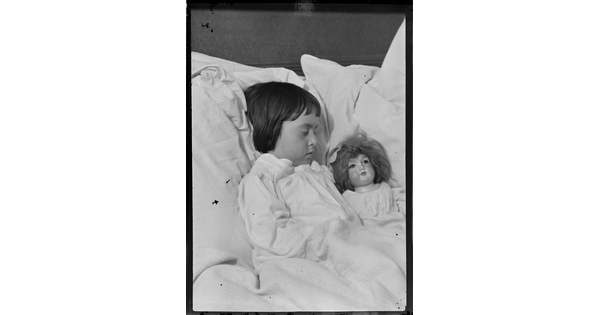 A black-and-white image of a young White girl, surrounded by pillows, sleeping next to a doll.