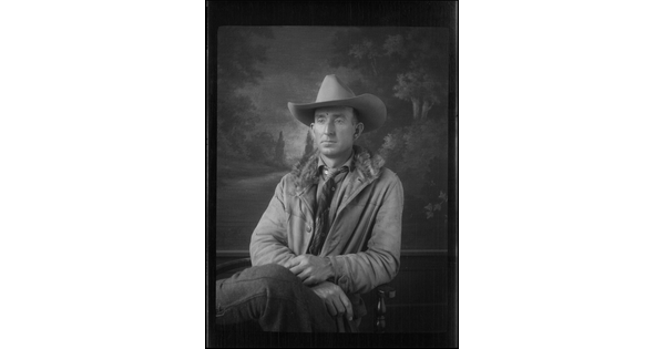 A black-and-white image of a seated White man wearing a cowboy hat, coat, and tie in front of a painted landscape.