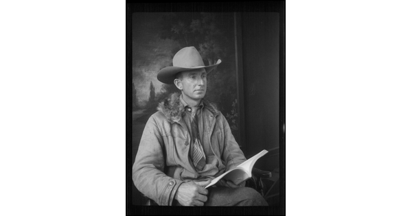 A black-and-white image of a seated White man wearing a cowboy hat, coat, and tie, holding an open book on his lap.