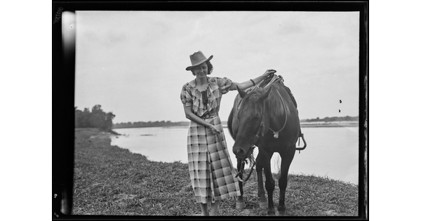 A black-and-white image of a White woman wearing a cowboy hat and plaid dress holding the reins and saddle of the horse next to her.