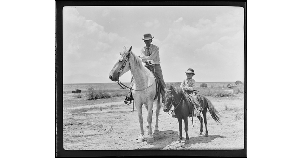 A black-and-white image of a White man wearing a cowboy hat on horseback and, next to him, a White child wearing a cowboy hat on a pony.