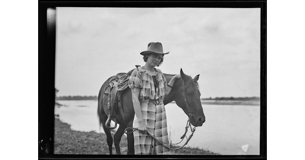 A black-and-white image of a White woman wearing a cowboy hat and plaid dress holding the reins of the horse standing next to her.
