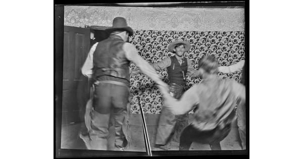 A black-and-white image of White cowboys holding hands dancing in a circle inside a room with bold patterned wallpaper.