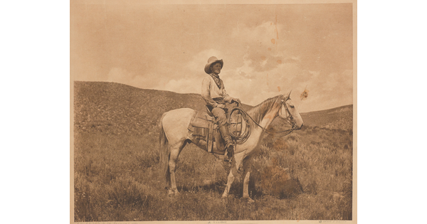 A sepia-toned photograph of a cowboy on a horse standing in a hilly plain.