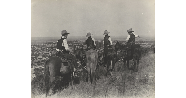 A black-and-white photograph of four cowboys on horseback, viewed from behind, looking over plains in the distance.