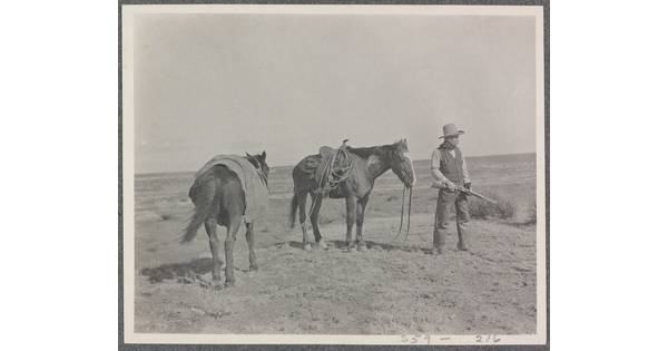 A black-and-white photograph of two saddled horses and a man, holding a rifle, standing on a grassy plain.