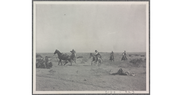 A black-and-white photograph of a group of men on horseback and one man lying on the ground.