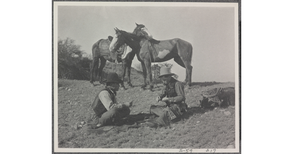 A black-and-white photograph of two cowboys playing a card game on the ground, while their horses stand nearby.