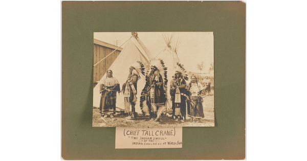 A sepia-toned photograph mounted on green board of a group of Native Americans in full regalia in front of tipis.