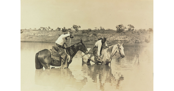 A sepia-toned photograph of two cowboys riding their horses through a river.