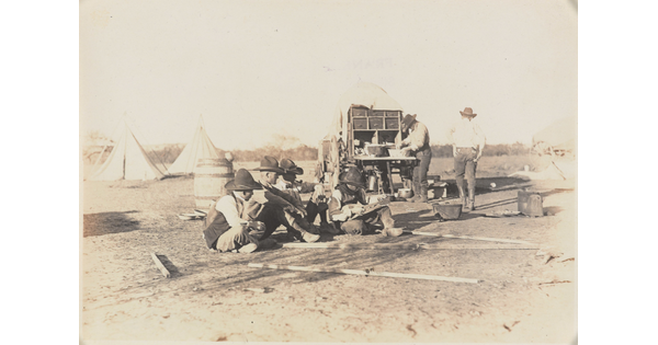 A black-and-white photograph of a group of cowboys, several seated on the ground, one walking, and one preparing food at a wagon; tipis in the background.