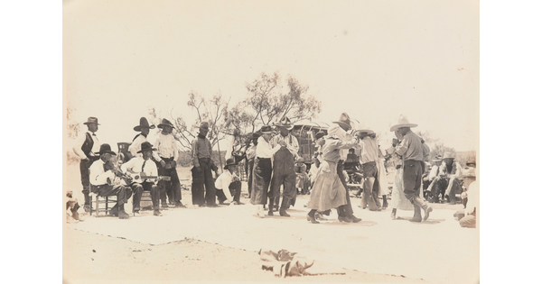 A black-and-white photograph of a group of people outdoors, most wearing cowboy hats, dancing as a band plays.