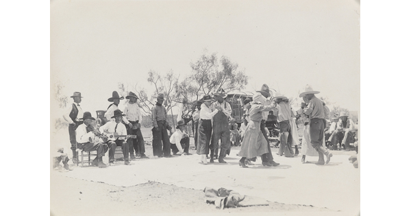 A black-and-white photograph of a group of people outdoors, most wearing cowboy hats, dancing as a band plays.