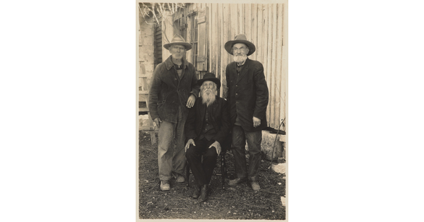 A black-and-white photograph of three men, two standing and one sitting, outside of a wood house, all wearing coats and hats.